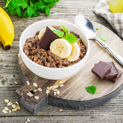 Chocolate oatmeal for breakfast with slices of a ripe banana and pieces of bitter good chocolate in a white ceramic bowl on a wooden background in a horizontal position. Example of a healthy die