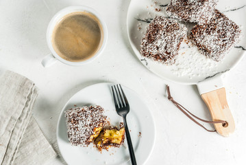 Australian food. Traditional dessert Lamington - pieces of biscuit in dark chocolate, sprinkled with coconut powder chips. On a marble plate, white table. With coffee mug. Copy space top view