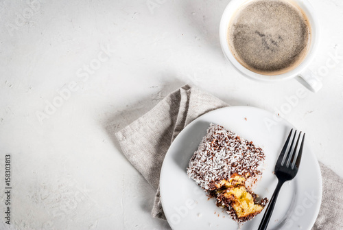 Australian food. Traditional dessert Lamington - pieces of biscuit in dark chocolate, sprinkled with coconut powder chips. On a marble plate, white table. With coffee mug. Copy space top view