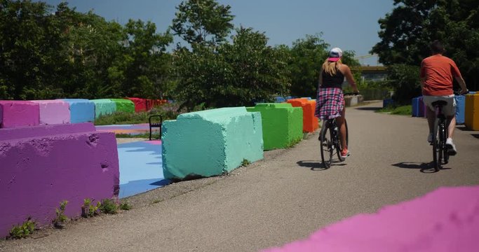 Two bicyclists pedal away from camera on a trail in Pittsburgh's South Side past colorful safety barriers on a sunny summer day.  	
