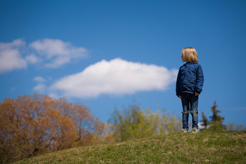 Portrait of cute blond kid boy standing on a hill on blue sky background on sunny spring day. Happy child walking in the park. Lifestyle.