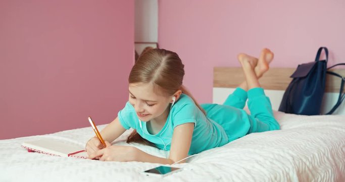 Student doing homework in exercise book. Child lying on bed in bedroom and listening music in headphones