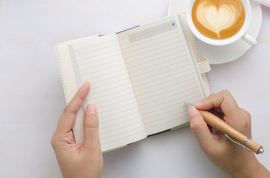 Woman hands with pen writing on notebook and coffee cup on white wooden table.