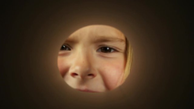 The inquisitive girl approaches the camera peering through the crack. A view through a round hole on a funny beautiful girl. Front view of little girl looking through a round opening.