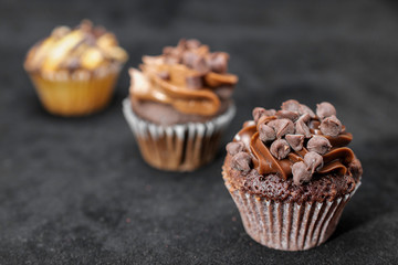 Three chocolate cupcakes garnished with chocolate chips and chocolated frosting on lined up across the frame, on black background, from left to right