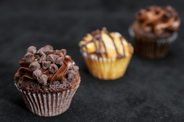 Three chocolate cupcakes garnished with chocolate chips and chocolate frosting on lined up across the frame, on black background, from right to left