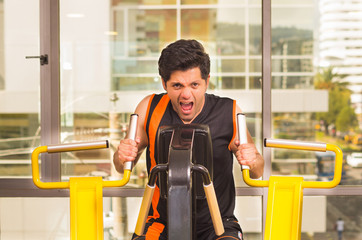 Handsome young man working on fitness machine at gym and screaming at same time