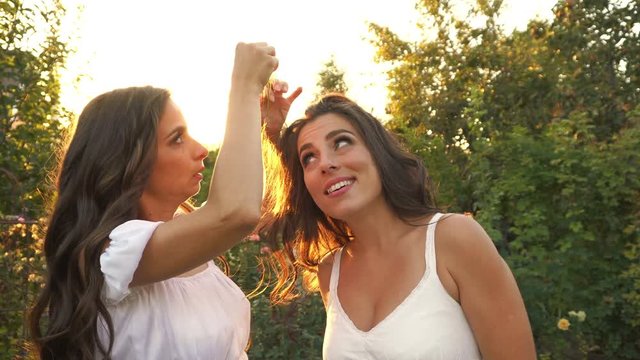 Two sisters in white dresses in the garden at sunset. The girl takes the hairpin out of her friend's hair. Family relationships.