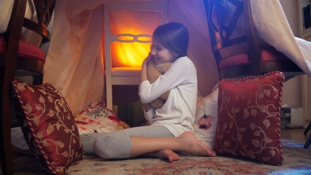 4k dolly video of cute girl in pajamas playing with teddy bear in selfmade tent at evening
