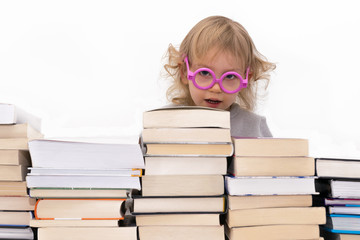 Three year old girl in pink glasses looking over stack of books. Isolated on white.