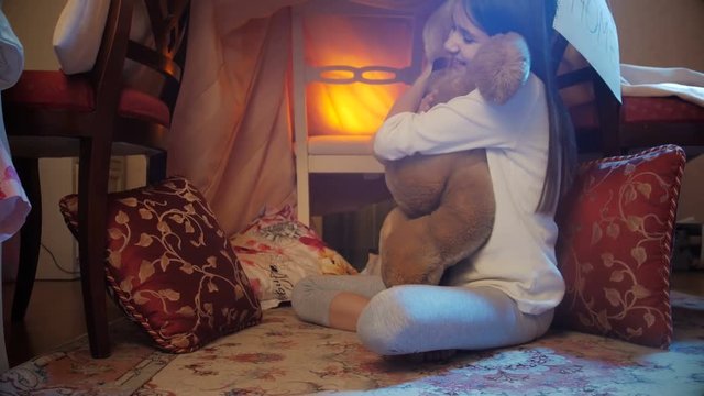 Dolly footage of smiling girl in pajamas sitting in tepee tent at bedroom and hugging teddy bear