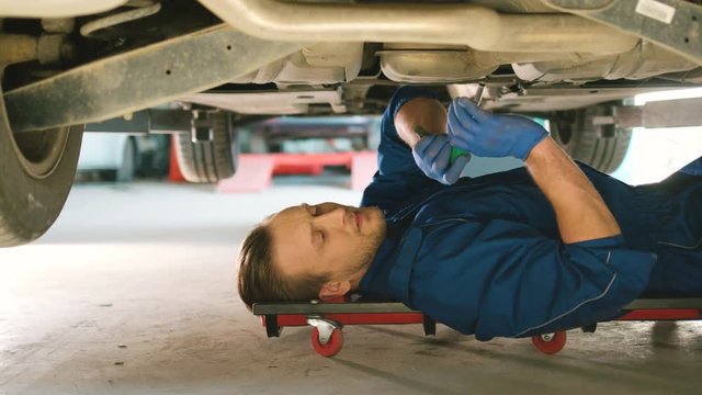 Side view of caucasian young auto mechanic man fastening the screws into the car bottom on autoservice background. Indoor.