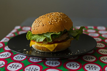 Homemade appetizing cheeseburger with melted cheese, lettuce leaf, cutlet, tomato in a toasted bun with sesame on a black plate on a red tablecloth with apples pattern on a gray background.