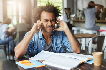 Hipster dark-skinned Afro American male with curly dark hair wearing denim shirt sitting at noisy cafeteria trying to hear voice over smartphone having bad conncetion working with books and writing
