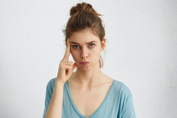 Serious attractive blue-eyed woman with hair knot wearing casual clothes holding finger on her temple having thoughtful expression. Female student having clever look isolated over white background