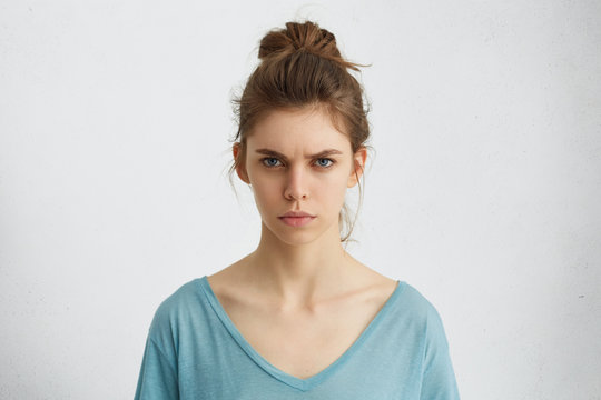 Portrait of outraged young woman with oval face, blue eyes and hair bun wearing blue casual sweater frowning her eyebrows being displeased with something. Scowling pretty female isolated over white