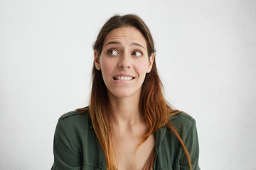 Fototapeta premium Indoor portrait of shy pleasant-looking female looking embarrassed aside biting her lower lip wanting to say something, but not having courage to do it. Woman feeling her guilt looking confused
