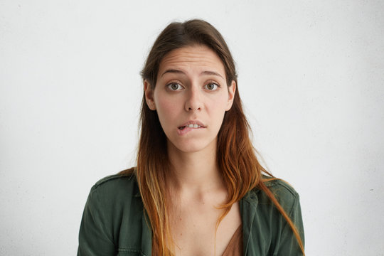 Horizontal portrait of puzzled woman with long face and straight dyed hair wearing green jacket looking with big opened eyes into camera biting her lower lip having some doubts and uncertainty