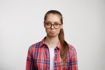 Portrait of offended female student with fair hair and pony tail wearing eyeglasses and checked shirt twisting her lips being upset to recieve bad mark. Young female blowing her cheeks with offence