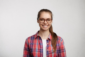 Cute female with green eyes and long eyelashes in big round eyeglasses and red checked shirt looking elegant having pleasant broad smile isolated over white background. Beauty and youth concept