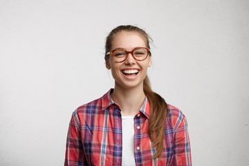 Adorable female in round spectacles and red checked shirt having fair hair tied in pony tail...