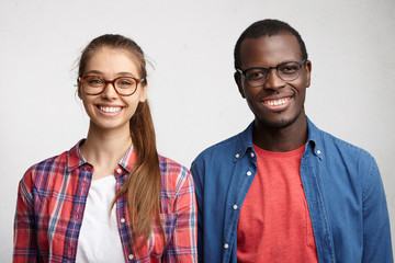 Portrait of interracial couple smiling with pleasure standing shoulder to shoulder. Cute Caucasian woman in glasses and checked shirt standing near her dark-skinned friend being happy to meet - Powered by Adobe