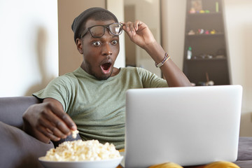 Horrified Afro American man dressed casually sitting at home watching horror films on computer...