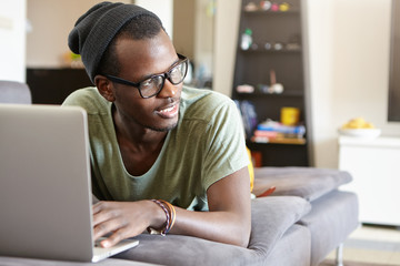 Fototapeta na wymiar Carefree hipster black guy in stylish hat and eyeglasses lying at cozy couch using laptop watching films and listening music isolated over home interior having rest during weekends having pensive look