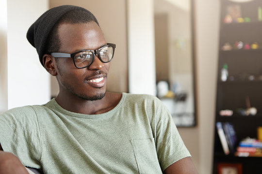 Dark-skinned student in stylish hat, eyeglasses and casual T-shirt relaxing after classes at home sitting at cozy sofa isolated over home interior thinking about his day. Relaxation and rest concept