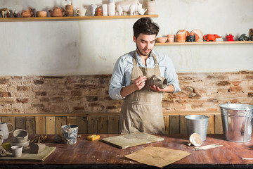 potter, workshop, ceramics art concept - smiling young brunette ceramist with raw product at workplace, male's hands holding the clay cup and tools, craftsman stand at a workplace behind a table