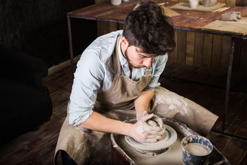 pottery, workshop, ceramics art concept - man working on potter's wheel with raw clay with hands, a male brunette sculpt a utensils near wooden table with tools, master in apron and a shirt, top view