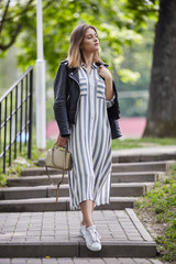 Young beautiful girl in stylish streetwear black leather jacket long striped dress white sneakers and with a fashionable bag strolling in summer city park