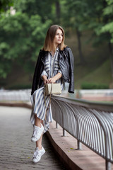 Young beautiful girl in stylish streetwear black leather jacket long striped dress white sneakers and with a fashionable bag strolling in summer city park