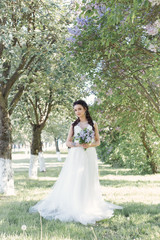 Fototapeta na wymiar Tender beautiful cute girl bride in a white air dress with a bouquet of lilacs in her hands walking through the park on a sunny spring day. Photo in gentle colors