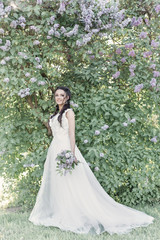 Obraz na płótnie Canvas Tender beautiful cute girl bride in a white air dress with a bouquet of lilacs in her hands walking through the park on a sunny spring day. Photo in gentle colors