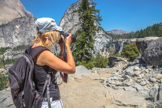 Travel photographer with professional camera takes shot of popular Nevada Falls on Merced River in Yosemite National Park. Woman mountain hiker in Yosemite waterfalls summit overlook, California, USA.