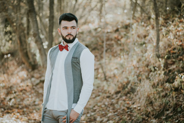 Portrait of stylish groom man in white shirt, gray waistcoat and red bow-tie at the wedding in nature forest. Selective focus. Toned image