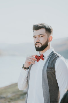 Portrait of stylish groom man in white shirt, gray waistcoat and red bow-tie at the wedding in nature forest and seacoast. Selective focus. Toned image