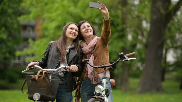 Funny Female Friends Holding Their Bikes are Taking Selfie Using Smartphone. Two Girls are Grimacing While Photographing Themselves by Mobile Phone in the City Park in Spring.