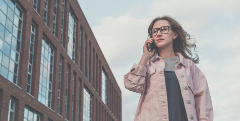 Portrait of young woman in glasses standing outdoors and talking on cellphone.Girl is walking along city street