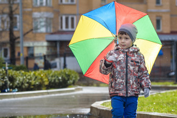 Obraz premium Little boy playing in rainy summer park. Child with colorful rainbow umbrella, waterproof coat and boots jumping in puddle and mud in the rain. Kid walking in autumn shower Outdoor fun by any weather