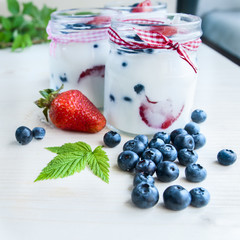 Three jars of yogurt with blueberries and strawberries, square. Berries of blueberries and strawberries are scattered on the table. Cooking, healthy food, food packaging
