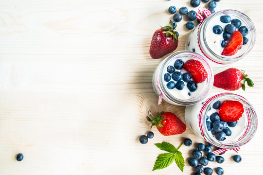Three jars of yogurt with blueberries and strawberries, square. Berries of blueberries and strawberries are scattered on the table. Cooking, healthy food, food packaging
