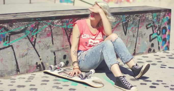 Young woman waiting at a skate park for a friend sitting against a low graffiti covered wall with her skateboard shielding her eyes from the hot summer sun.