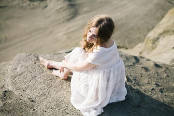 Красивая девушка в белом платье с длинными русыми волосами сидит на песке The beautiful girl in a white dress with a long fair hair sits on sand