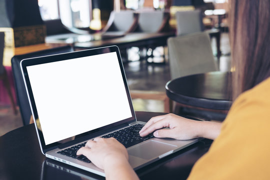 Mockup image of a woman using laptop with blank white screen on wooden table in modern cafe