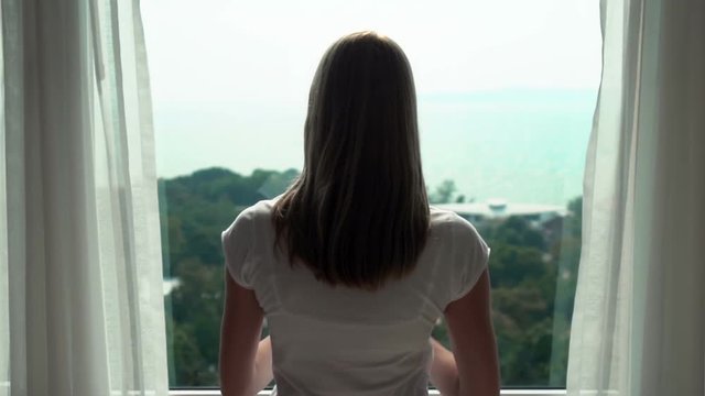 Woman in white t-shirt unveiling curtains and looking out of window. Enjoying the sea view outside