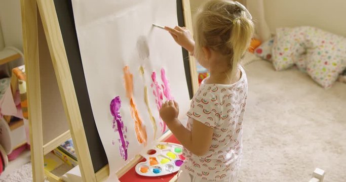 Little blond girl standing painting at an easel creating a colorful abstract picture in her playroom viewed close up from behind with a view of the painting and her palette.