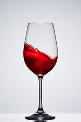 Moving red wine in the elegant wineglass standing against light background with reflection.