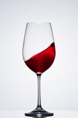 Moving red wine in the pure fragile wineglass standing against light background with reflection.